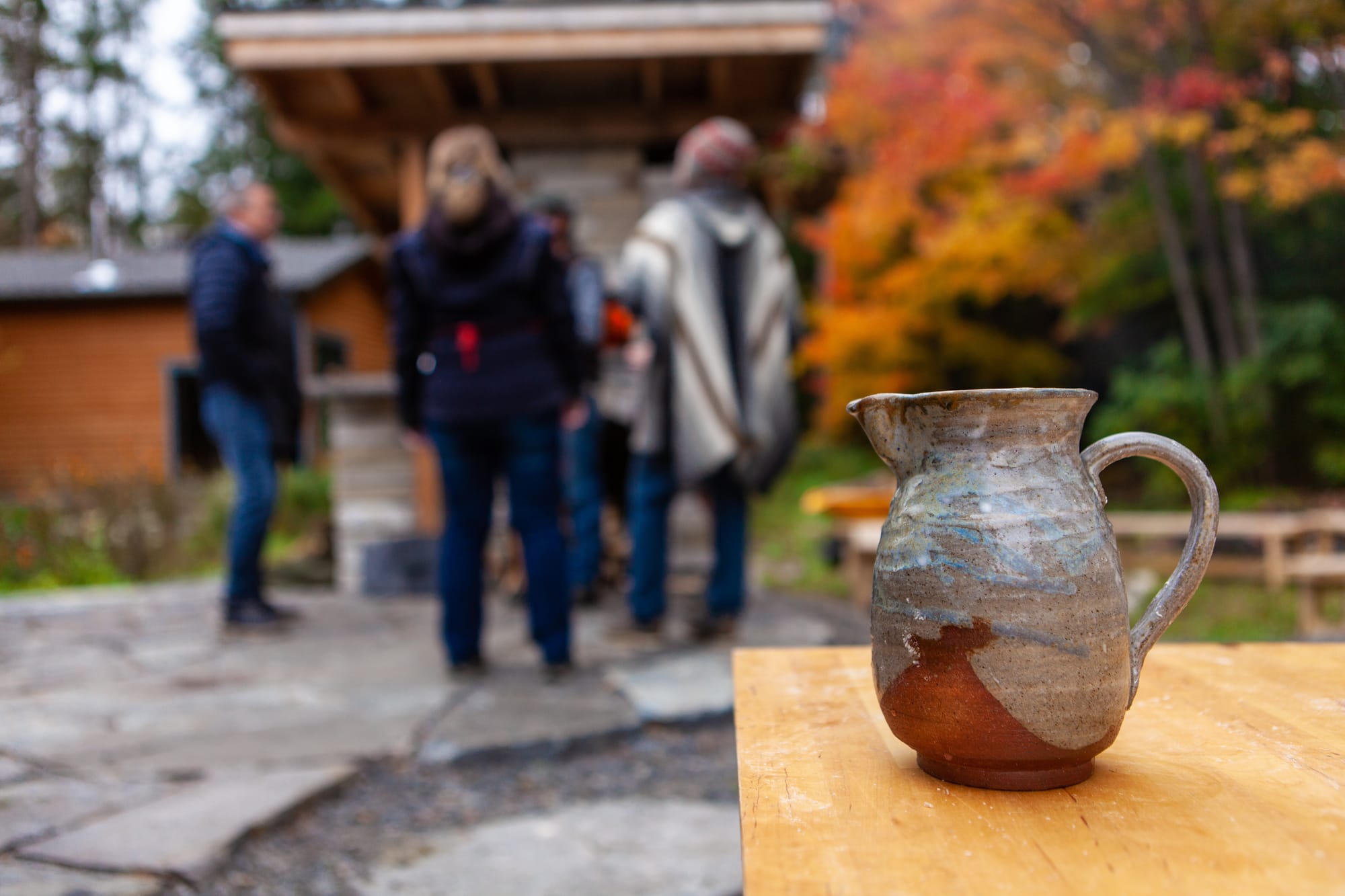 Outdoor setting in autumn. Pottery mug of tea in foreground with friends standing in a circle chatting in background.